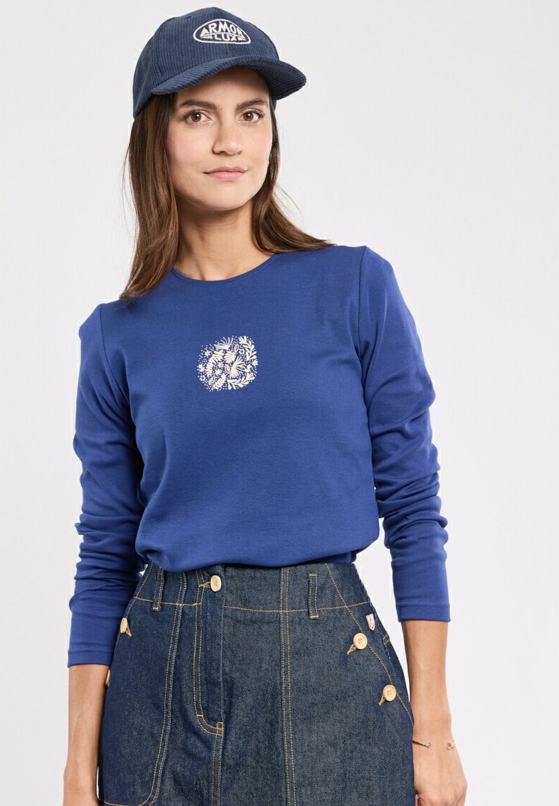 Niebieska koszula z długim rękawem i okrągłym dekoltem, z białą grafiką kwiatową. W komplecie ze spódnicą dżinsową podkreśloną guzikami i kieszeniami po bokach.