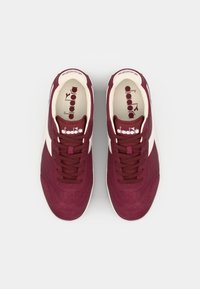 Tenisi bordo cu un brâu din suede texturat, accente crem și șireturi plate. Caracteristici includ un guler căptușit și un interior cu marcă, cu logo.