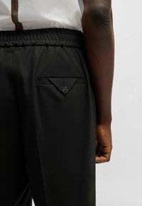 Black trousers with a textured waist and an angled patch pocket secured by a button on the back, smooth fabric with a tailored look.