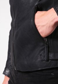 Giacca in pelle nera con chiusura a zip, tasca laterale con zip e texture liscia. I dettagli includono cuciture e un accento con nastro laterale.