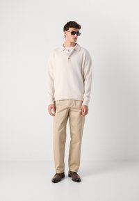 Man in beige coltrui met kraag, lichtbruine broek, bruine nette schoenen en donkere zonnebril die tegen een witte achtergrond staat.