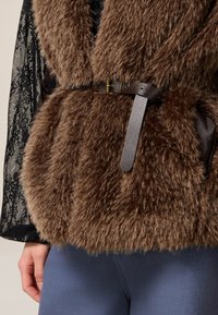 Bordeaux nep bont gilet met een gestructureerde uitstraling, voorzien van een donkerbruine nep leren riem en een gouden gesp. Gelaagd over een zwart kanten top.