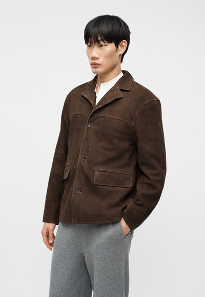 Νέος άνδρας που φοράει ένα σκούρο καφέ σατέν μπουφάν με τρία κουμπιά και γκρι παντελόνια, στέκεται με το δεξί χέρι στην τσέπη μπροστά σε απλό φόντο.