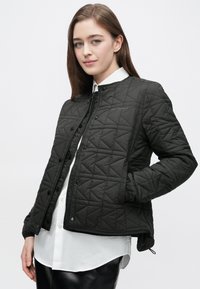 Jachetă neagră matlasată cu model în formă de diamant, nasturi cu capse și decolteu rotund, asortată cu o cămașă albă cu nasturi dedesubt.