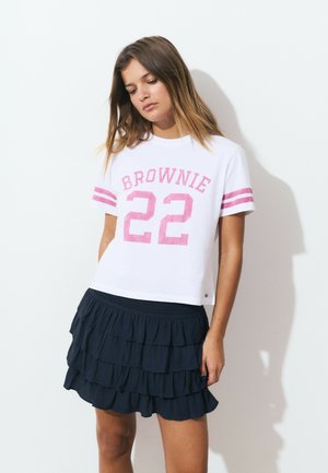 Jeune femme portant un T-shirt blanc avec des rayures roses et le texte "BROWNIE 22", associé à une jupe noire à volants superposés, sur fond blanc.