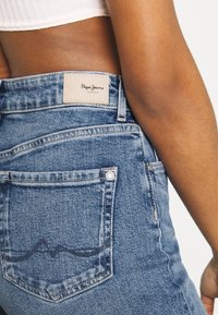 Vaqueros de mezclilla azul con cintura alta, con una etiqueta marrón, bolsillos traseros y un sutil detalle de costura en el bolsillo trasero.