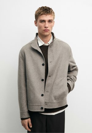 Junger Mann mit kurzen blonden Haaren, der eine graue, geknöpfte Jacke über einem braunen Pullover und einem weißen Hemd mit Kragen trägt, steht vor einem schlichten weißen Hintergrund.