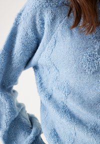 Hellblau gestrickter Pullover mit strukturierten Bobble-Akzenten, lockerer Passform und gerippten Bündchen. Der Stoff wirkt weich und gemütlich.