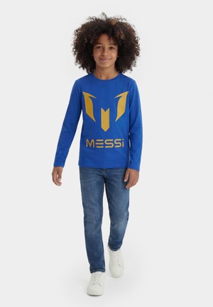 Barn med krøllet hår, der går, iført en blå langærmet skjorte med et gyldent "Messi"-logo, blå jeans og hvide sneakers på en hvid baggrund.