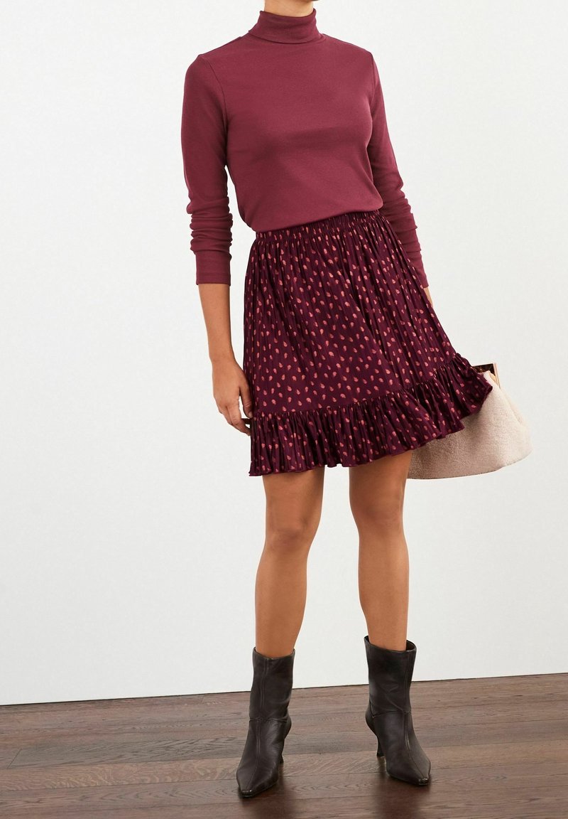 Femme portant un col roulé bordeaux à manches longues et une jupe plissée assortie avec un ourlet à volants, des bottines noires et tenant un sac beige.