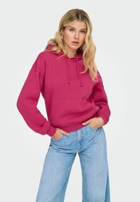 Femme aux longs cheveux blonds portant un hoodie magenta et un jean large bleu clair, se tenant avec les mains dans les poches contre un fond blanc.