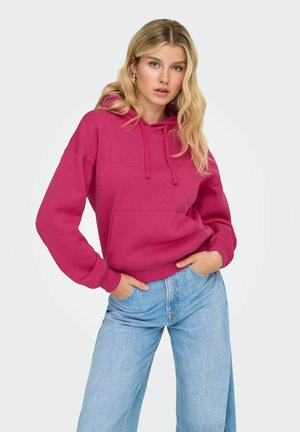 Vrouw met lang blond haar, gekleed in een magenta hoodie en lichtblauwe wijde jeans, staande met handen in de zakken tegen een witte achtergrond.