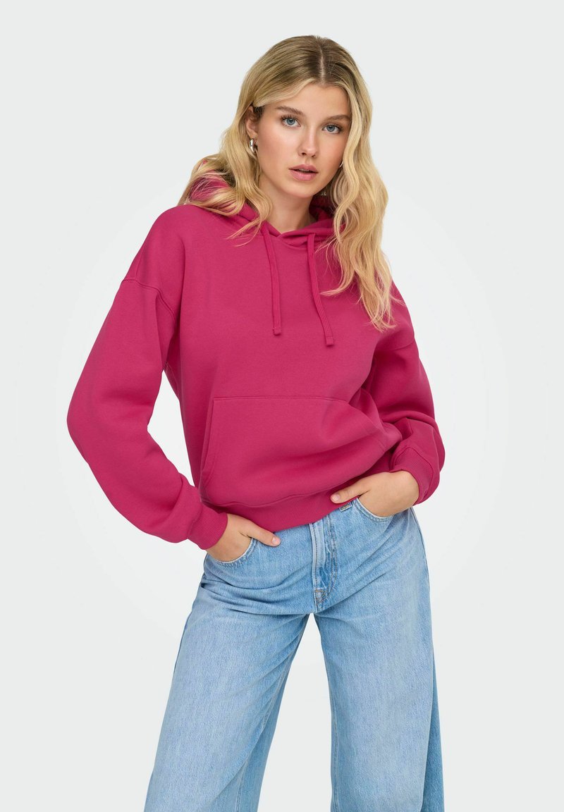 Femme aux longs cheveux blonds portant un hoodie magenta et un jean large bleu clair, se tenant avec les mains dans les poches contre un fond blanc.