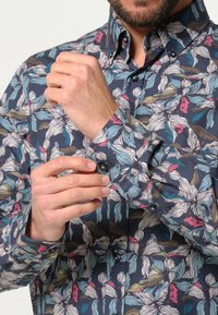 Een button-up shirt met een donkerblauwe achtergrond en een bloemenpatroon in het wit, roze en blauw. Lange mouwen met knoopmanchetten.