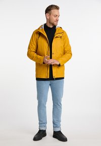 Giacca impermeabile gialla con cappuccio, zip frontale e chiusura a bottoni. Maglione nero sotto, jeans azzurri chiari e scarpe nere.
