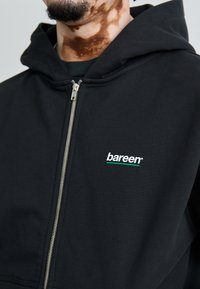 Sort zip-up hoodie lavet af bomuldsblanding, med et logo i hvidt og grønt på venstre brystområde, blød tekstur og justerbar hætte.