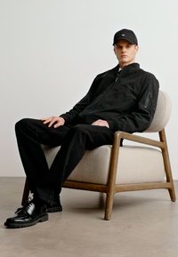 Černá zipová bunda s bočním kapsou, černé kalhoty a černé boty. Sedí na světlé čalouněné židli s dřevěným rámem.