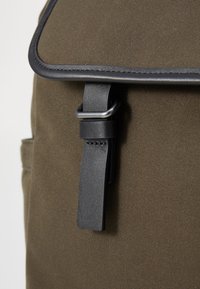 Sac à dos en toile vert olive avec une sangle en cuir noir et une boucle en métal. Présente des bords cousus et une surface texturée.