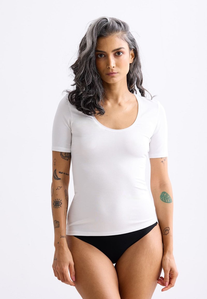 White short-sleeve shirt with a scoop neckline, paired with black bottom. Model features visible tattoos on arms. Smooth texture.