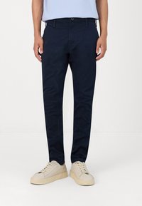 Chinos azul marino hechos de una mezcla de algodón, con un corte ajustado, bucles para cinturón y bolsillos laterales, combinados con zapatillas de colores claros.