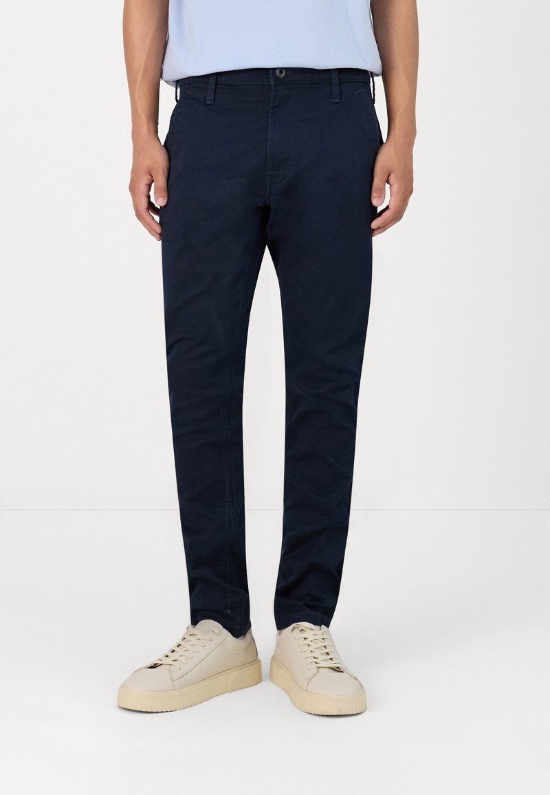 Chinos azul marino hechos de una mezcla de algodón, con un corte ajustado, bucles para cinturón y bolsillos laterales, combinados con zapatillas de colores claros.