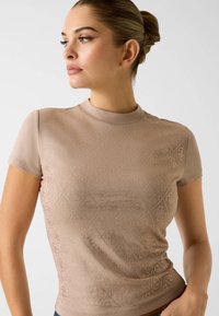 Krótki rękaw, dopasowany top w jasnobeżowym kolorze z fakturowanym wzorem diamentu i subtelnym połyskiem. Gładka tkanina, wysoki dekolt i minimalna liczba szwów.