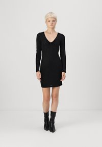 Vestido preto de manga longa com decote em V, apresentando um tecido texturizado. A barra fica acima do joelho, combinado com botas pretas até ao joelho.