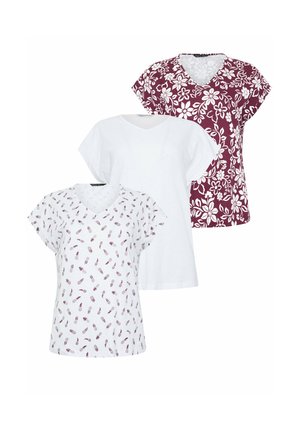 Frau mit lockigem Haar, die ein weißes V-Ausschnitt-T-Shirt mit kleinen Ananasdruck trägt, blaue hochgekrempelte Jeans und weiße Turnschuhe, und an einer Wand lehnt.