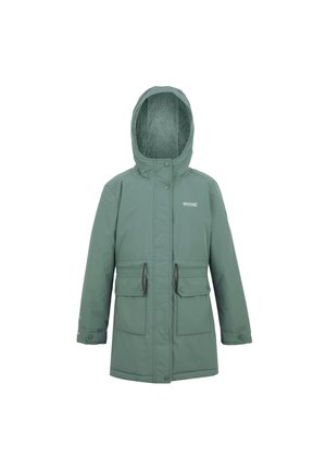 Młoda dziewczyna o długich blond włosach, ubrana w turkusową kurtkę i beżowe spodnie, uśmiechnięta, stojąca na tle jednolitego, jasnego tła.