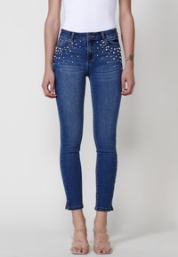 Skinny blue denim jeans featuring pearl embellishments along the front pockets and a cropped hem with a raw edge.
