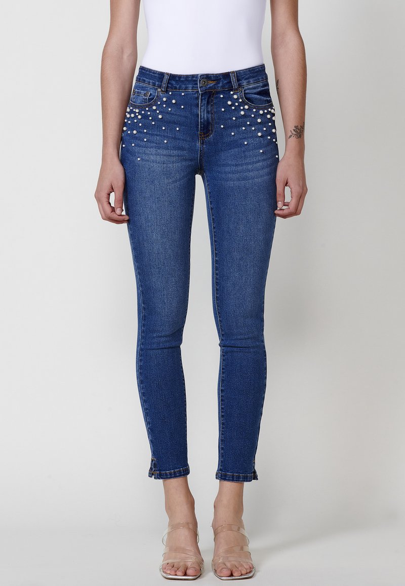 Skinny blue denim jeans featuring pearl embellishments along the front pockets and a cropped hem with a raw edge.