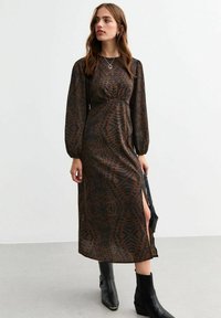 Brązowa midi sukienka w wzory z długimi rękawami, z podkreśloną talią i bocznym rozcięciem. Noszona z czarnymi botkami za kostkę. Gładka tekstura materiału.