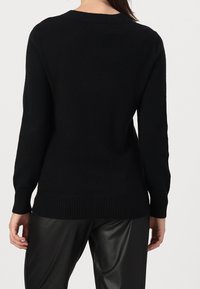 Woman wearing a black long-sleeve sweater and black leather pants, shown from the back on a light background.