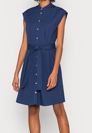 Kvinde iført en ærmeløs, mørkeblå kjole med knapper foran og bånd i taljen, stående mod en ensartet, lys baggrund.