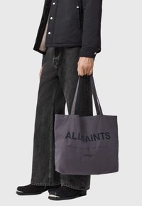 Pilka medvilninė pirkinių rankinė su „ALLSAINTS“ logotipu, ilgos rankenos. Dėvima su juodais džinsais ir tamsiais batais. Paprastas dizainas ir kasdienė tekstūra.