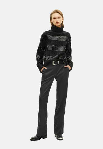 Giovane vestito con un maglione a collo alto nero a motivi, pantaloni grigio scuro su misura, cintura nera e stivali neri, in piedi con le mani in tasca.