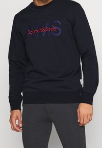 Námořnická mikina s výšivkou "AMS" a "Scotch & Soda" v červené a modré barvě, s žebrovanými manžetami a spodním lemem. Hladká tkanina, ležérní design.