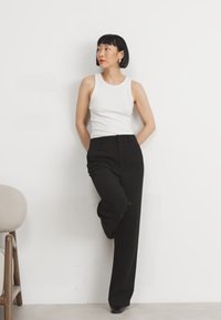 Svarte striped bukser med en avsmalnet design, paret med en hvit ermeløs topp. Modellen poserer mot en ensfarget bakgrunn.