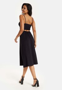 Black midi dress with thin straps, gathered back, cut-out sides, and a flowing silhouette. Worn with black block-heeled sandals.