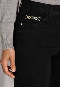 Hand rustend nabij de zwarte broekzak met gouden decoratieve sluiting, persoon in een lichtgrijze lange mouwen trui.