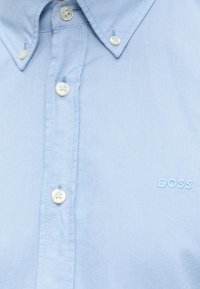 Hellblaues Hemd mit weißen Knöpfen und "BOSS" in passendem blauem Faden auf der Brust gestickt.
