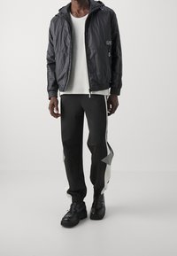 Hombre vestido con chaqueta ligera con capucha negra, camisa blanca, pantalones deportivos en blanco y negro, y mocasines negros contra un fondo liso.