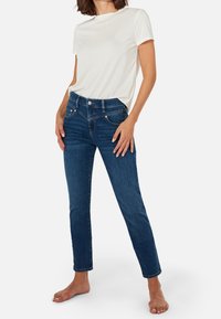 Jeans de mezclilla azul con un ajuste ajustado y cintura media, que cuentan con cinco bolsillos y un cierre visible con botón y cremallera. Combinados con una camiseta blanca lisa.
