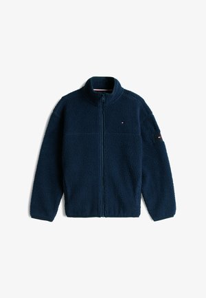 Ragazzo in piedi che indossa una giacca in pile blu navy, una t-shirt bianca, jeans jogger blu e sneakers bianche con strisce rosse e nere.