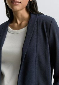 Cardigan navy con un tessuto a maglia testurizzata, design aperto davanti e una morbida camicia a coste color crema sotto, evidenziando colori e materiali a contrasto.