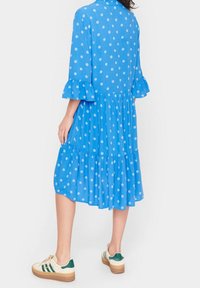 Blå klänning med prickigt mönster, rynkade ärmar och flerlagers kjol, tillsammans med krämfärgade sneakers med gröna detaljer. Slät tygtextur.