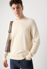 Cream-colored knit sweater with a round neckline and ribbed cuffs, featuring a small, square logo patch on the chest. Worn with dark jeans.