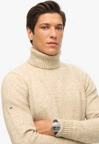 Suéter de cuello alto beige con cuello acanalado, patrón de tejido texturizado y mangas ajustadas. Reloj plateado con correa negra en la muñeca.