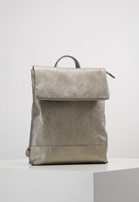 Metallinen hopeinen nahkarepulla, jossa on läppäkiinnitys ja yläkäsikäsi, seisoo suorana valkoisella jalustalla yksinkertaista taustaa vasten.