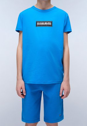 Chłopiec ubrany w jasnoniebieską koszulkę z krótkim rękawem z logo "Napapijri geographic" i pasujące niebieskie szorty, stojący na jednolitym tle.